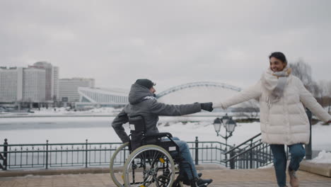 Glückliche-Muslimische-Frau-Und-Ihr-Behinderter-Freund-Im-Rollstuhl,-Der-Sich-Im-Winter-In-Der-Stadt-An-Den-Händen-Hält