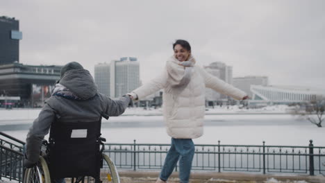 Happy-Muslim-Woman-And-Her-Disable-Friend-In-Wheelchair-Spinning-Holding-Hands-In-City-In-Winter-1
