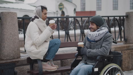 Muslim-Woman-And-Her-Disabled-Friend-In-Wheelchair-Drinking-Takeaway-Coffe-On-A-Bench-In-City-In-Winter