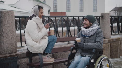 Muslimische-Frau-Und-Ihr-Behinderter-Freund-Im-Rollstuhl-Trinken-Kaffee-Zum-Mitnehmen-Auf-Einer-Bank-In-Der-Stadt-Im-Winter-1