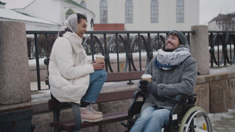 Muslimische-Frau-Und-Ihr-Behinderter-Freund-Im-Rollstuhl-Trinken-Kaffee-Zum-Mitnehmen-Auf-Einer-Bank-In-Der-Stadt-Im-Winter-2