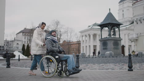 Muslimische-Frau,-Die-Ihren-Behinderten-Freund-Im-Rollstuhl-Im-Winter-Bei-Einem-Spaziergang-Durch-Die-Stadt-Mitnimmt