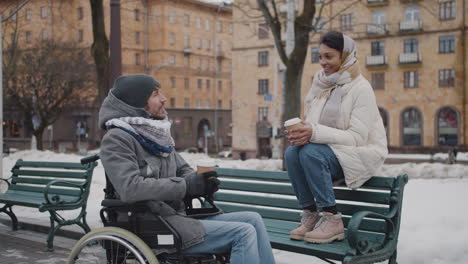 Muslimische-Frau-Und-Ihr-Behinderter-Freund-Im-Rollstuhl-Trinken-Kaffee-Zum-Mitnehmen-Auf-Einer-Bank-In-Der-Stadt-Im-Winter-4