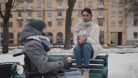 Muslim-Woman-And-Her-Disabled-Friend-In-Wheelchair-Drinking-Takeaway-Coffe-On-A-Bench-In-City-In-Winter-5