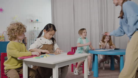 Female-Teacher-Giving-Play-Dough-To-Her-Pupils-Sitting-At-Desk-In-Montessori-School