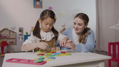 Little-Girl-In-A-Montessori-School-Playing-With-Shapes-Stacking-While-Teacher-Helping-Her