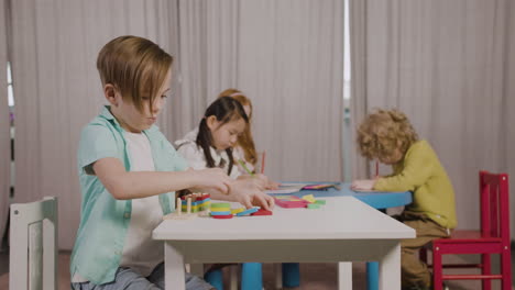 Kleiner-Junge-In-Einer-Montessori-Schule,-Der-Mit-Formen-Spielt,-Die-Sich-Stapeln,-Während-Seine-Klassenkameraden-Zeichnen