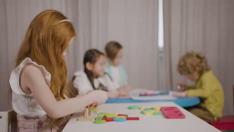 Ingwer-Kleines-Mädchen-In-Einer-Montessori-Schule,-Das-Mit-Formen-Spielt,-Die-Sich-Stapeln,-Während-Ihre-Klassenkameraden-Zeichnen