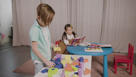 Kleiner-Junge-In-Einer-Montessori-Schule,-Der-Mit-Schaumstoffbausteinen-Spielt,-Während-Ein-Kleines-Mädchen-Am-Schreibtisch-Sitzt-Und-In-Einem-Buch-Blättert