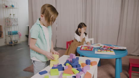 Kleiner-Junge-In-Einer-Montessori-Schule,-Der-Mit-Schaumstoffbausteinen-Spielt,-Während-Ein-Kleines-Mädchen-Am-Schreibtisch-Sitzt-Und-Zeichnet