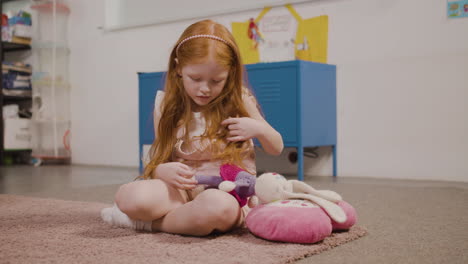 Redhead-Little-Girl-Picking-Up-Toy-Rabbit-While-Sitting-On-The-Floor-In-Classroom-In-A-Montessori-School