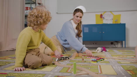 Der-Kleine-Blonde-Junge-Und-Sein-Lehrer-Spielen-Mit-Holzautos-Auf-Dem-Teppich-Im-Klassenzimmer-Einer-Montessori-Schule