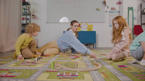 Little-Blond-Boy-And-Redhead-Little-Girl-Playing-With-Their-Teacher-With-Wooden-Cars-On-The-Carpet-In-Classroom-In-A-Montessori-School