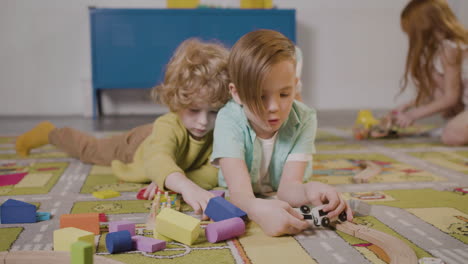Front-View-Of-Two-Little-Boys-Playing-With-Wooden-Pieces-And-Car-Lying-On-A-Carpet-In-A-Montessori-School-Class