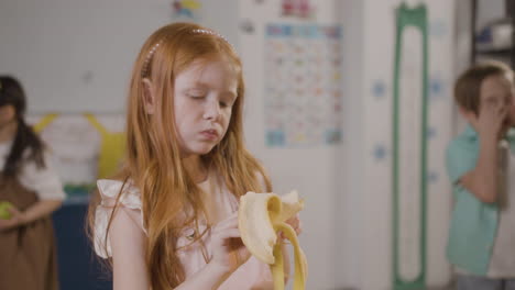 Rothaariges-Mädchen,-Das-Eine-Banane-Im-Klassenzimmer-In-Einer-Montessori-Schule-Isst
