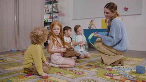 Vier-Kinder-Sitzen-Auf-Einem-Teppich,-Während-Ihr-Lehrer-Ihnen-Hilft,-Lange-Luftballons-In-Einer-Montessori-Schule-Aufzublasen