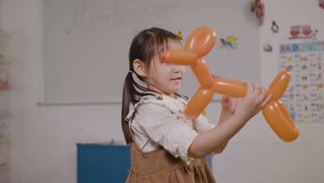 Kleines-Mädchen,-Das-Mit-Einem-Langen-Ballon-In-Form-Eines-Hundes-Spielt,-Während-Es-Im-Klassenzimmer-In-Einer-Montessori-Schule-In-Die-Kamera-Blickt