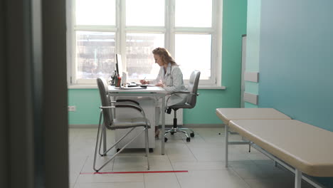 Smiling-Female-Doctor-Writing-On-Paper-While-Sitting-At-Desk-In-Her-Consulting-Room