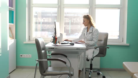 Ärztin,-Die-Am-Computer-Arbeitet,-Während-Sie-In-Ihrem-Sprechzimmer-Am-Schreibtisch-Sitzt
