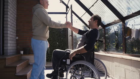 Behinderter-Mann-Im-Rollstuhl-Und-Sein-Freund-Grüßen-Mit-Händedruck,-Umarmen-Sich-Und-Machen-Zu-Hause-Fauststoß