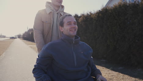 Young-Man-Walking-While-Pushing-His-Disabled-Friend-In-Wheelchair-And-Laughing-Together