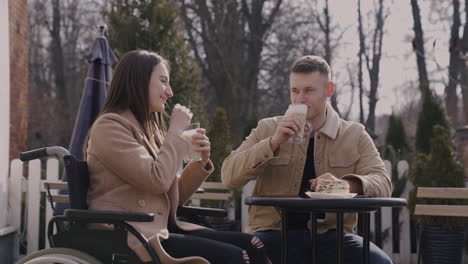 Disabled-Girl-In-Wheelchair-And-Her-Male-Friend-Drinking-Milkshake-And-Talking-Together-While-Sitting-In-A-Bar-Terrace