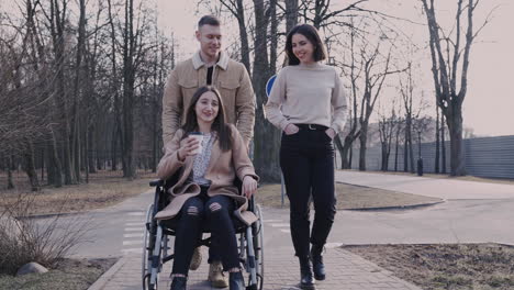 Front-View-Of-Woman-And-Man-Taking-A-Walk-With-Her-Disable-Friend-In-Wheelchair-Around-The-City