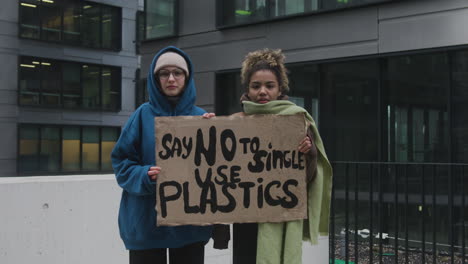 Zwei-Junge-Klimaaktivistinnen-Halten-Ein-Banner-Und-Protestieren-Gegen-Die-Einwegkunststoffe,-Während-Sie-In-Die-Kamera-Schauen