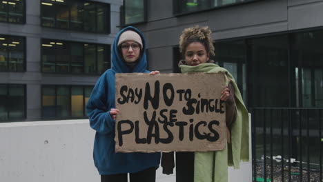Zwei-Junge-Klimaaktivistinnen-Halten-Ein-Banner-Hoch-Und-Protestieren-Gegen-Einwegkunststoffe,-Während-Sie-Auf-Kamera-1-Blicken