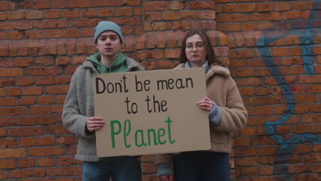 Young-Male-And-Female-Activists-Looking-At-Camera-Holding-A-Cardboard-Placard-During-A-Climate-Change-Protest