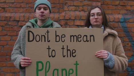 Close-Up-View-Of-Young-Male-And-Female-Activists-Looking-At-Camera-Holding-A-Cardboard-Placard-During-A-Climate-Change-Protest