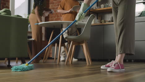 Close-Up-Of-An-Unrecognizable-Woman-Mopping-The-Floor-While-Her-Two-Roommates-Cleaning-The-Kitchen
