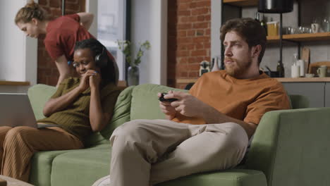 Afroamerican-Girl-Using-Laptop-And-Boy-Playing-Video-Games-Sitting-On-Sofa-While-Their-Roommate-Cleaning-The-House-1