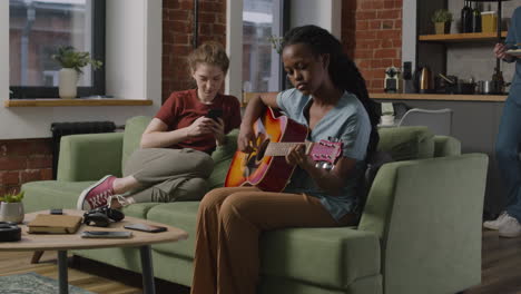 Mädchen,-Das-Gitarre-Spielt,-Sitzt-Auf-Dem-Sofa,-Während-Ihre-Mitbewohnerin-Das-Smartphone-Benutzt