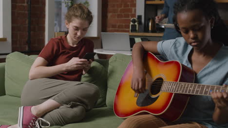 Girl-Playing-Guitar-Sitting-On-Sofa-While-Her-Female-Roommate-Using-The-Smartphone-1