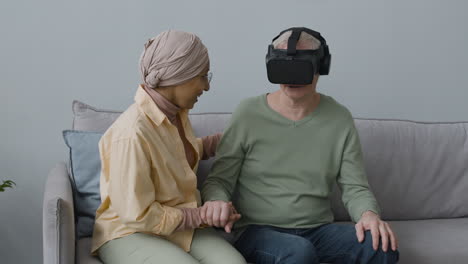Middle-Aged-Arabic-Woman-Helping-A-Senior-Man-With-Virtual-Reality-Glasses-While-Sitting-Together-On-Sofa-At-Home