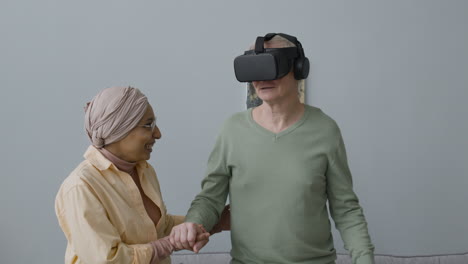 Mujer-árabe-De-Mediana-Edad-Ayudando-A-Un-Anciano-Con-Gafas-De-Realidad-Virtual-A-Levantarse-Del-Sofá-En-Casa