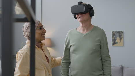 Senior-Man-Using-Virtual-Reality-Glasses-At-Home-While-An-Middle-Aged-Arabic-Woman-Helping-Him-To-Find-The-Right-Position-In-The-Room