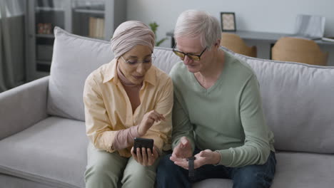 Middle-Aged-Arabic-Woman-Helping-A-Senior-Man-To-Sync-A-Smartwatch-To-Phone-While-Sitting-On-Sofa-At-Home