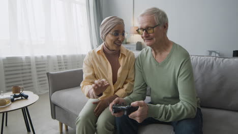 Middle-Aged-Arabic-Woman-Explaining-To-A-Senior-Man-How-To-Use-The-Game-Controller-While-Sitting-Together-On-Couch-At-Home