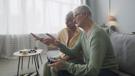 Middle-Aged-Arabic-Woman-Explaining-To-A-Senior-Man-How-To-Use-The-Game-Controller-While-Sitting-Together-On-Couch-At-Home-1