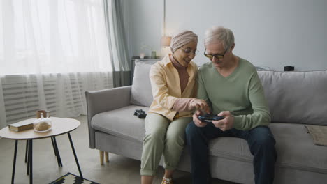 Middle-Aged-Arabic-Woman-Explaining-To-A-Senior-Man-How-To-Use-The-Game-Controller-While-Sitting-Together-On-Couch-At-Home-2