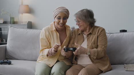 Middle-Aged-Arabic-Woman-Explaining-To-A-Senior-Lady-How-To-Use-The-Game-Controller-While-Sitting-Together-On-Couch-At-Home
