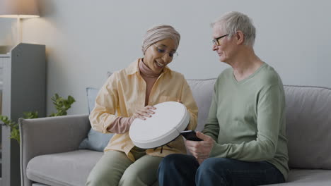 Middle-Aged-Arabic-Woman-Explaining-To-A-Senior-Man-How-To-Use-The-Robot-Vacuum-Cleaner-While-Sitting-Together-On-Couch-At-Home