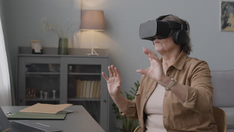 Senior-Woman-Using-Virtual-Reality-Headset-Glasses-And-Moving-Her-Hands-While-Sitting-On-Chair-In-A-Modern-Living-Room