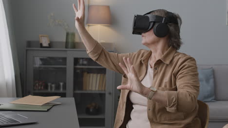 Senior-Woman-Using-Virtual-Reality-Headset-Glasses-And-Moving-Her-Hands-While-Sitting-On-Chair-In-A-Modern-Living-Room-1