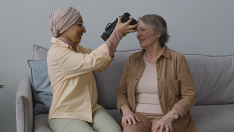 Arabische-Frau-Mittleren-Alters,-Die-Einer-älteren-Dame-Hilft,-Zu-Hause-Eine-Virtual-Reality-Headset-Brille-Zu-Verwenden