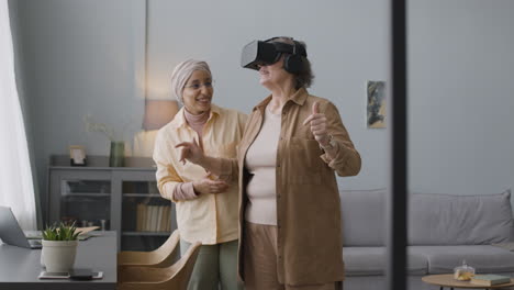 Middle-Aged-Arabic-Woman-Helping-A-Happy-Senior-Lady-To-Use-Virtual-Reality-Headset-Glasses-At-Home