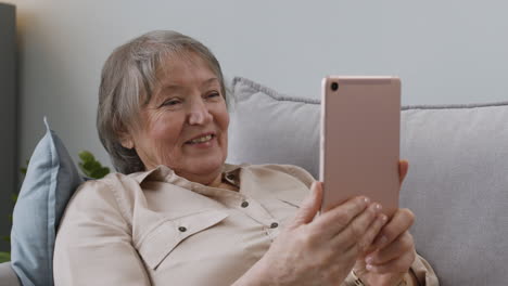 Glückliche-Seniorin,-Die-Einen-Videoanruf-Per-Tablet-Hat,-Während-Sie-Zu-Hause-Auf-Dem-Sofa-Liegt
