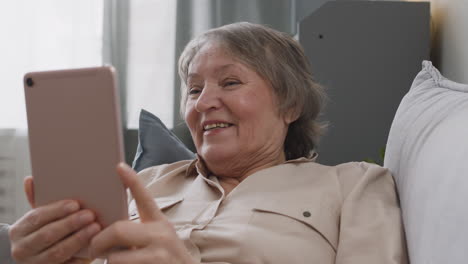 Glückliche-Seniorin,-Die-Einen-Videoanruf-Per-Tablet-Hat,-Während-Sie-Zu-Hause-Auf-Dem-Sofa-Liegt-1